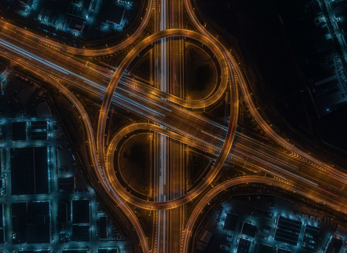 An ultra-crisp aerial night photograph of a sweeping highway interchange on the outskirts of a city, seen from directly overhead. Interlacing overpasses and looping ramps form an elegant, abstract pattern of curves and lines, delineated by continuous ribbons of white and red vehicle lights. Sodium-vapor and LED streetlights cast a refined mixture of warm amber and cool blue tones over the asphalt, creating subtle gradations of color and shadow. Surrounding industrial lots and darkened open spaces recede into a minimal, textured backdrop. The composition emphasizes symmetry and flowing movement, with every lane and barrier rendered in precise photographic detail, evoking a sophisticated, almost sculptural vision of contemporary infrastructure from above.